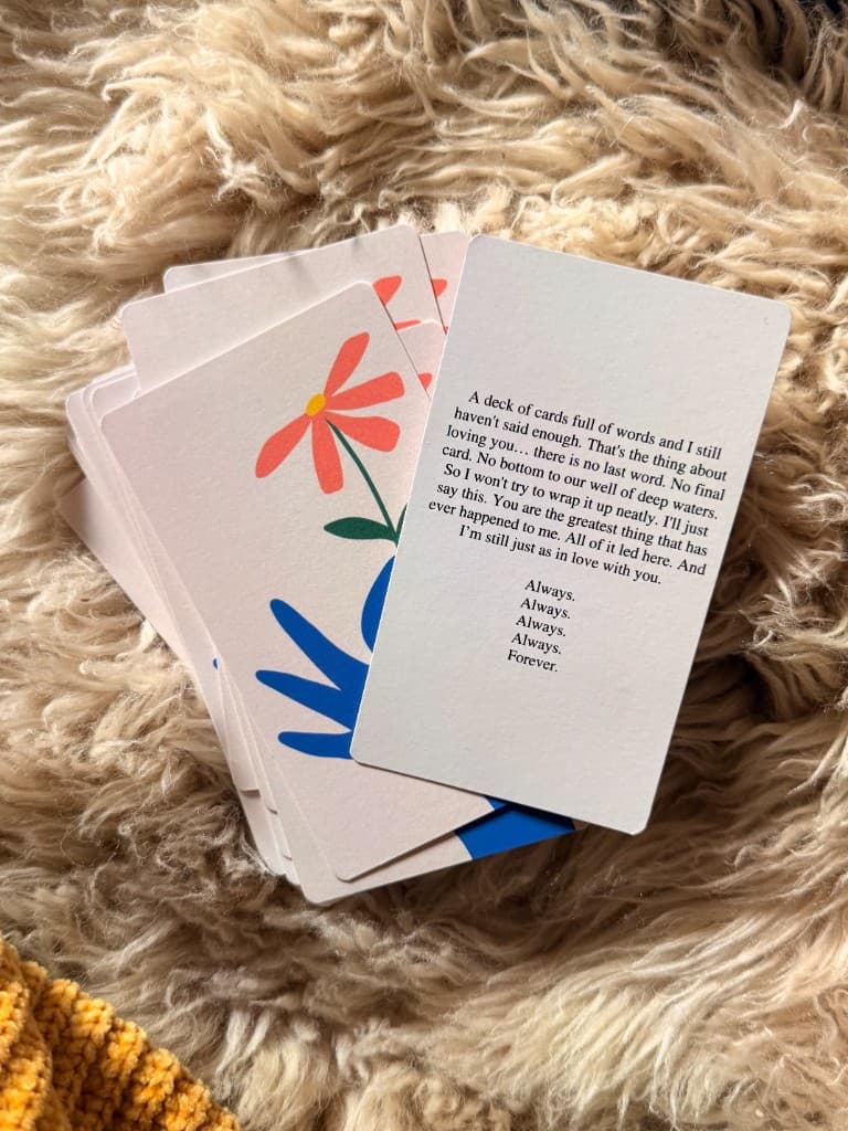 A fanned-out SayMore card deck resting on a soft cream blanket, with a handwritten card on top reading 'A deck of cards full of words and I still haven't said enough...'